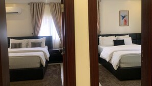 A hotel room with two beds, a dark headboard, a window with curtains, an air conditioning unit, and a painting on the wall. - Tennys Place Apartments GARKI (Abuja)