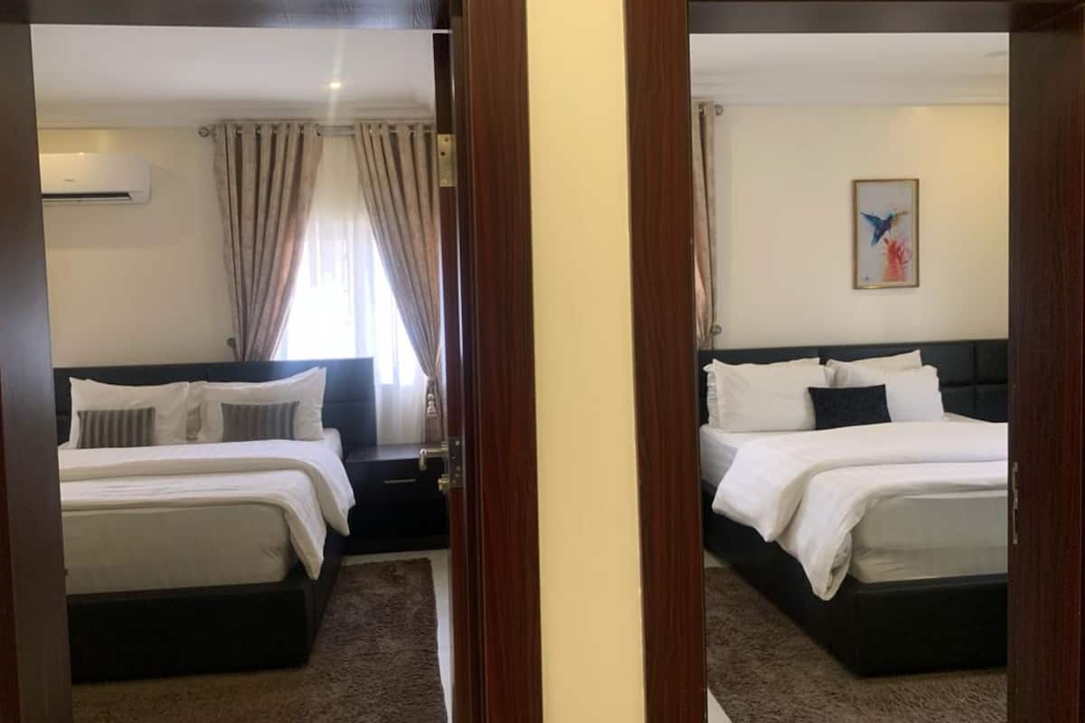 A hotel room with two beds, a dark headboard, a window with curtains, an air conditioning unit, and a painting on the wall.