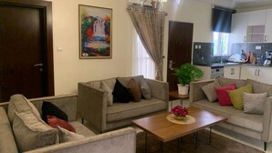 A living room with two sofas, a coffee table, a painting, and a kitchen in the background. - Tennys Place Apartments GARKI (Abuja)