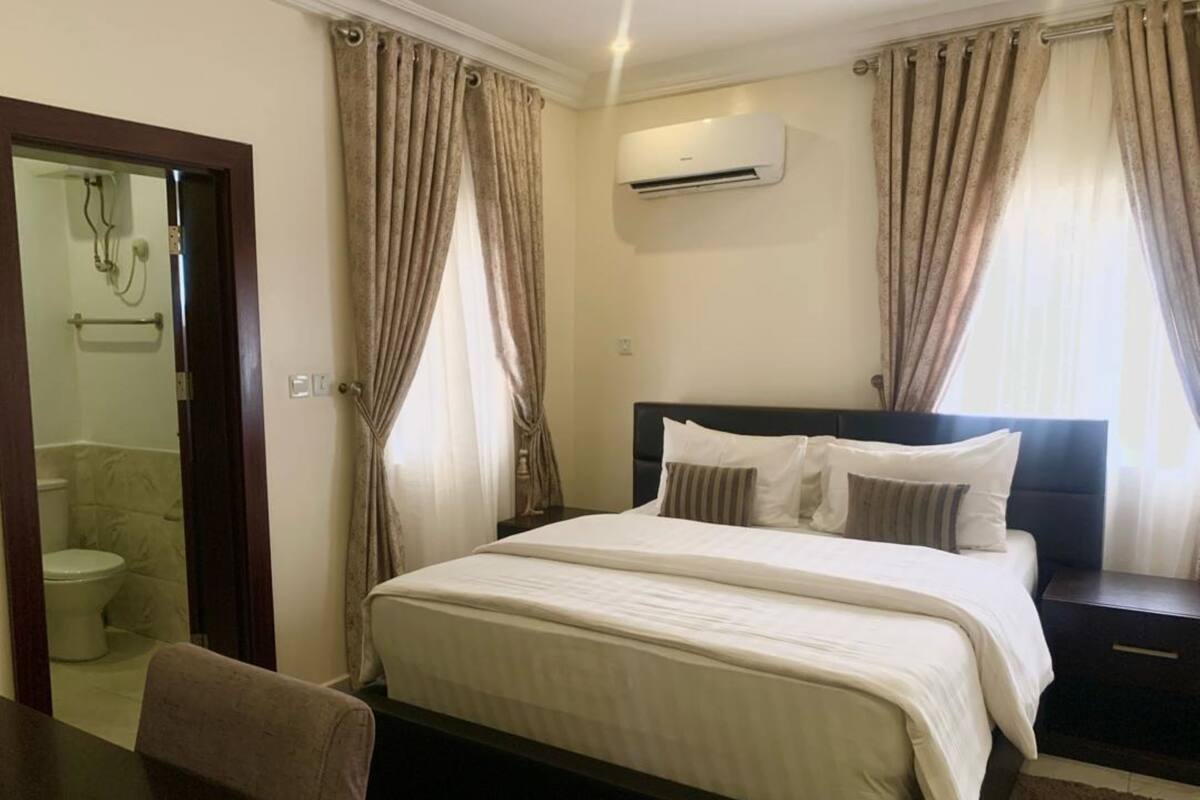 A hotel room with a bed, bedside tables, an air conditioning unit, and curtains.