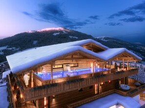 Un chalet en bois doté d’un jacuzzi et d’une piscine, entouré d’arbres et de montagnes couverts de neige.