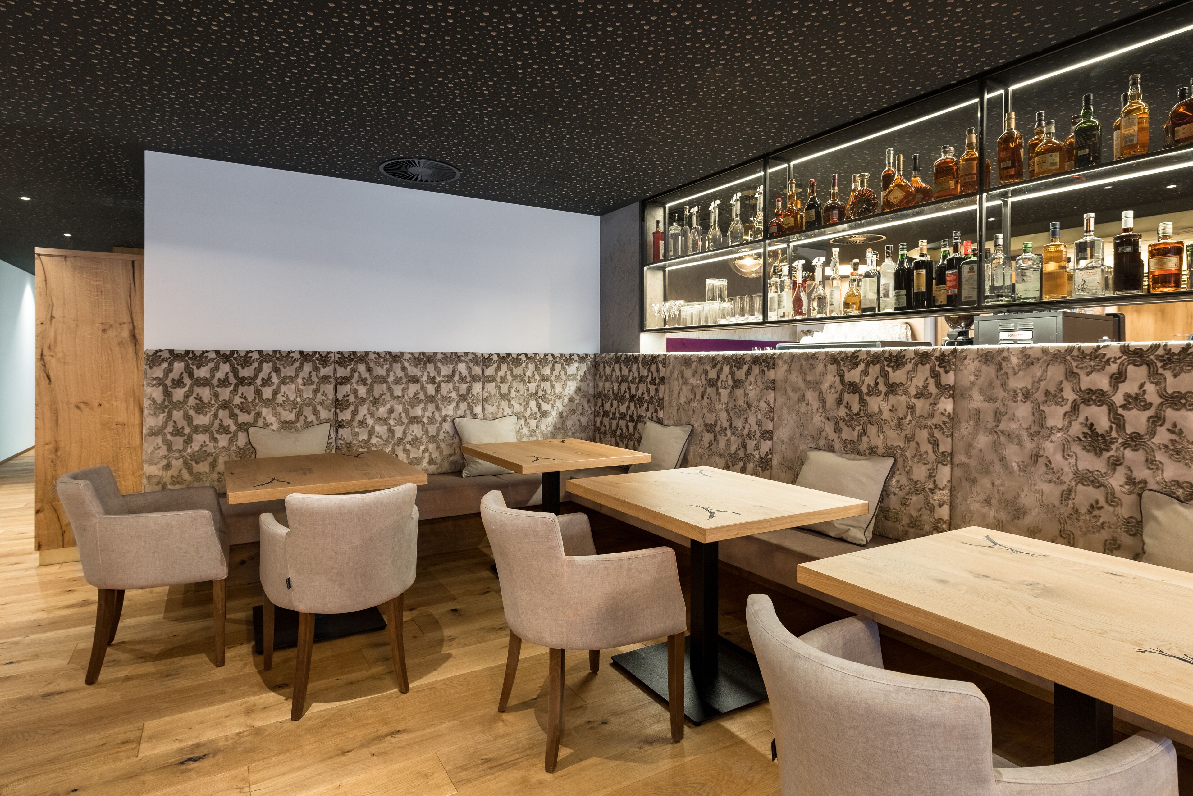 A modern bar area with wooden tables and chairs, a wall-mounted liquor display, and patterned wallpaper.