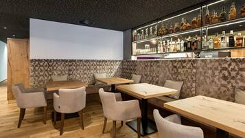 Uma área de bar moderna com mesas e cadeiras de madeira, um display de bebidas fixado na parede e papel de parede estampado.