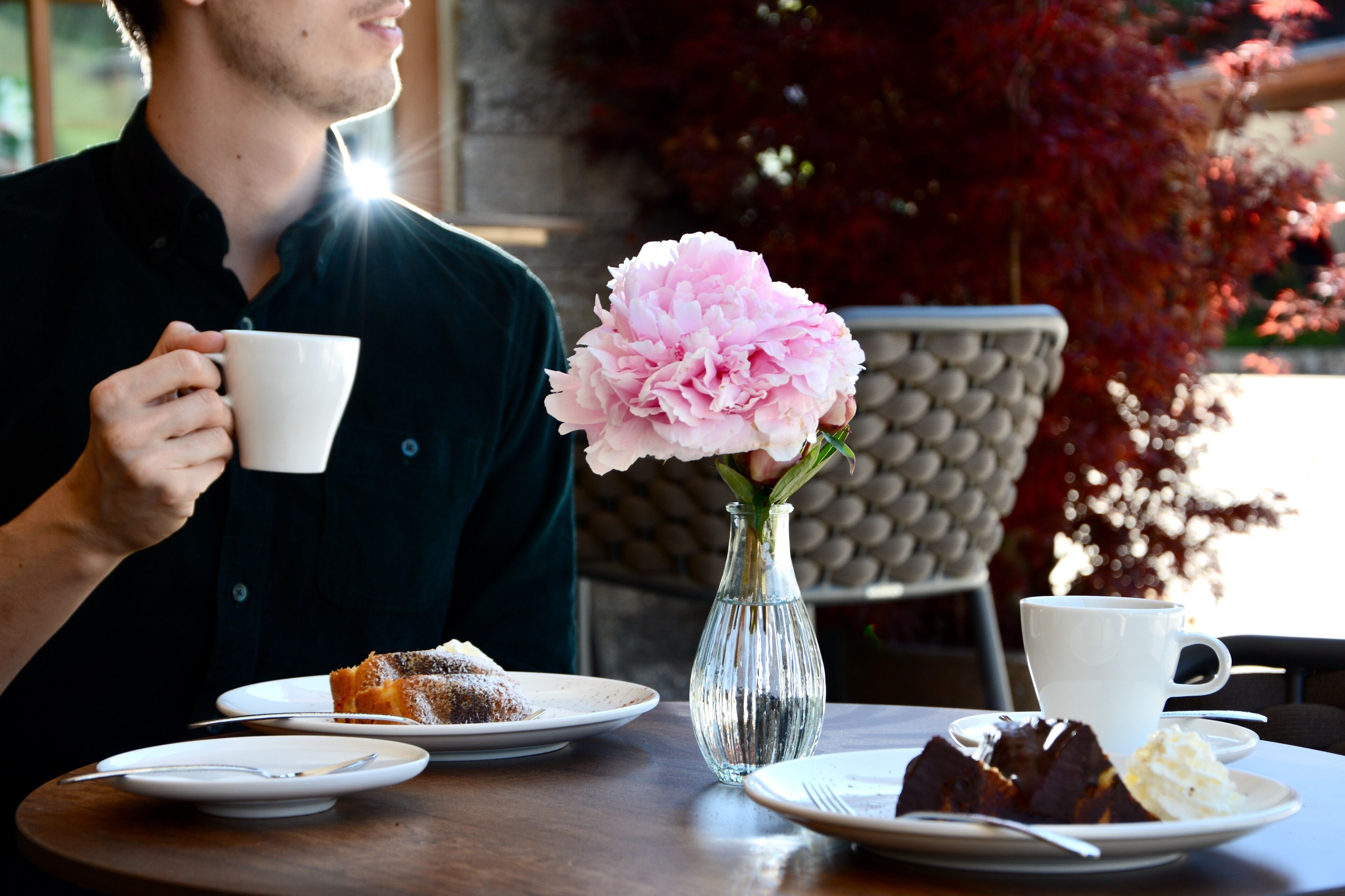 Une personne tenant une tasse blanche, assise à une table où se trouvent des assiettes de nourriture, un vase avec une fleur rose et une chaise, dans un café.
