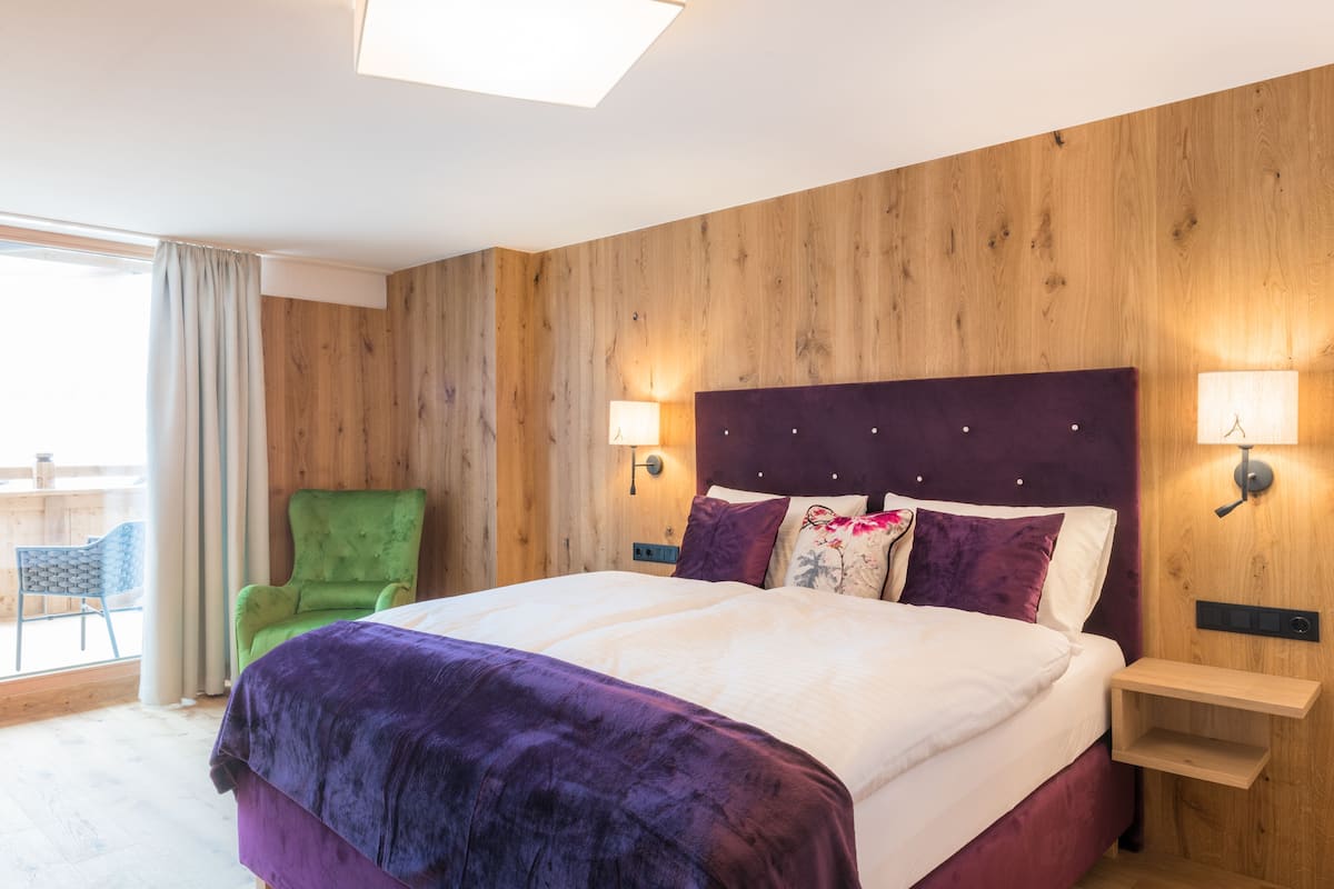 A hotel room with a wooden wall, a large bed with a purple bedspread, two bedside lamps, and a green chair.