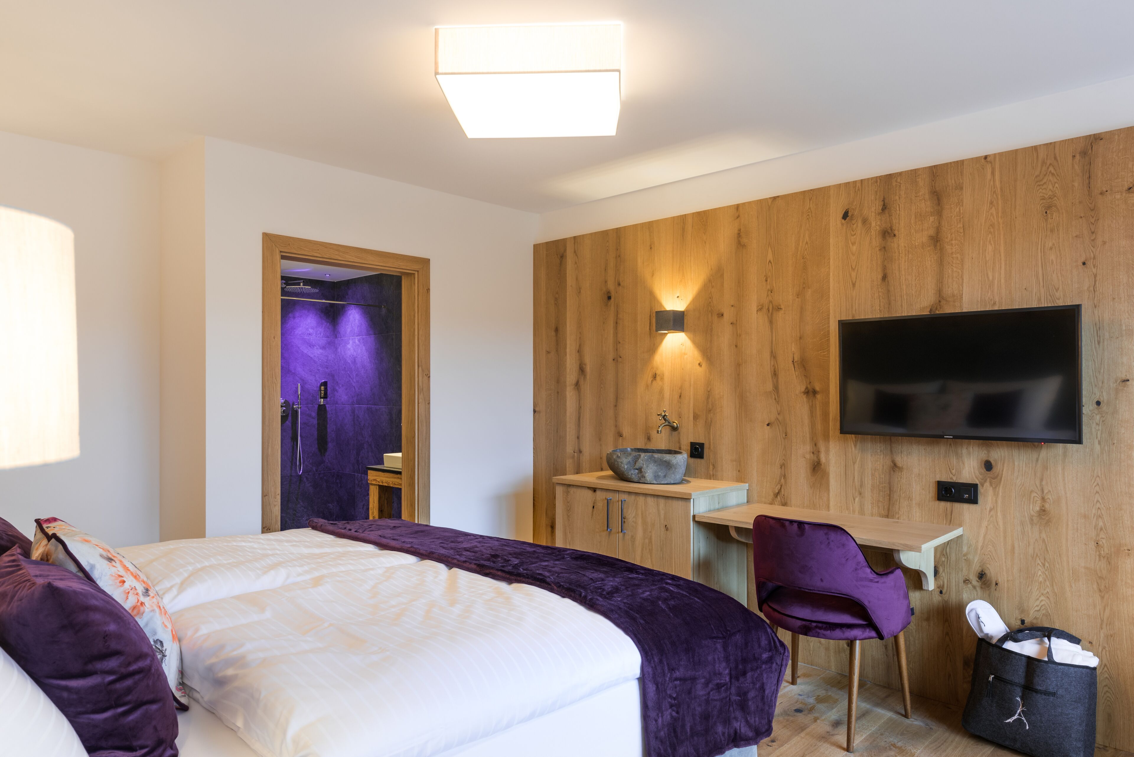 A modern hotel room with a bed, a desk, a chair, a television, and a bathroom with purple lighting.