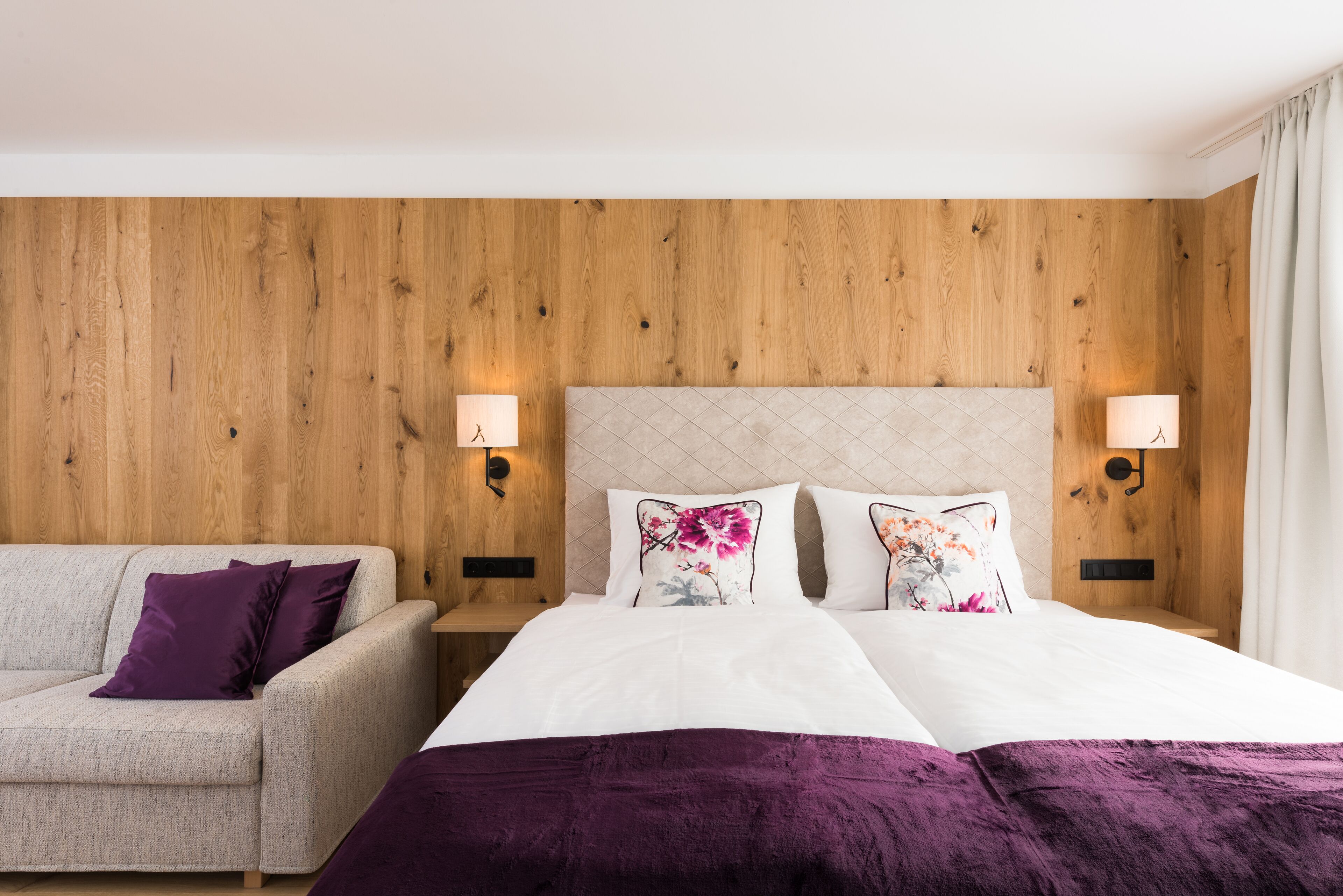 A hotel room with a wooden headboard, a bed with white linens and purple accents, a beige sofa, and two wall-mounted lamps.