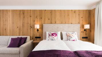 A hotel room with a wooden headboard, a bed with white linens and purple accents, a beige sofa, and two wall-mounted lamps.