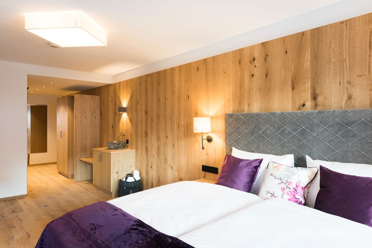 A modern hotel room with a wooden wall, a bed with a grey headboard, and a nightstand with a lamp.