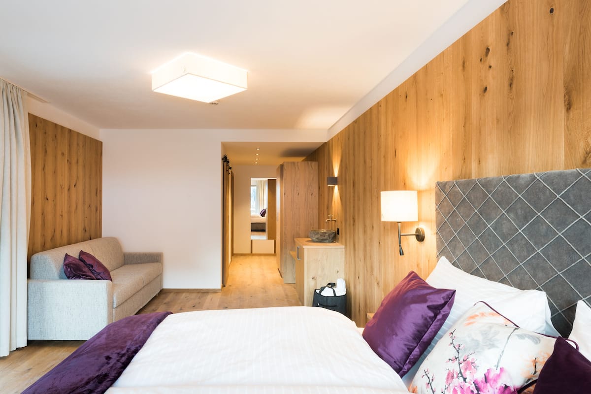 A hotel room with a bed, a sofa, a wooden wall, and a bathroom visible through an open door.