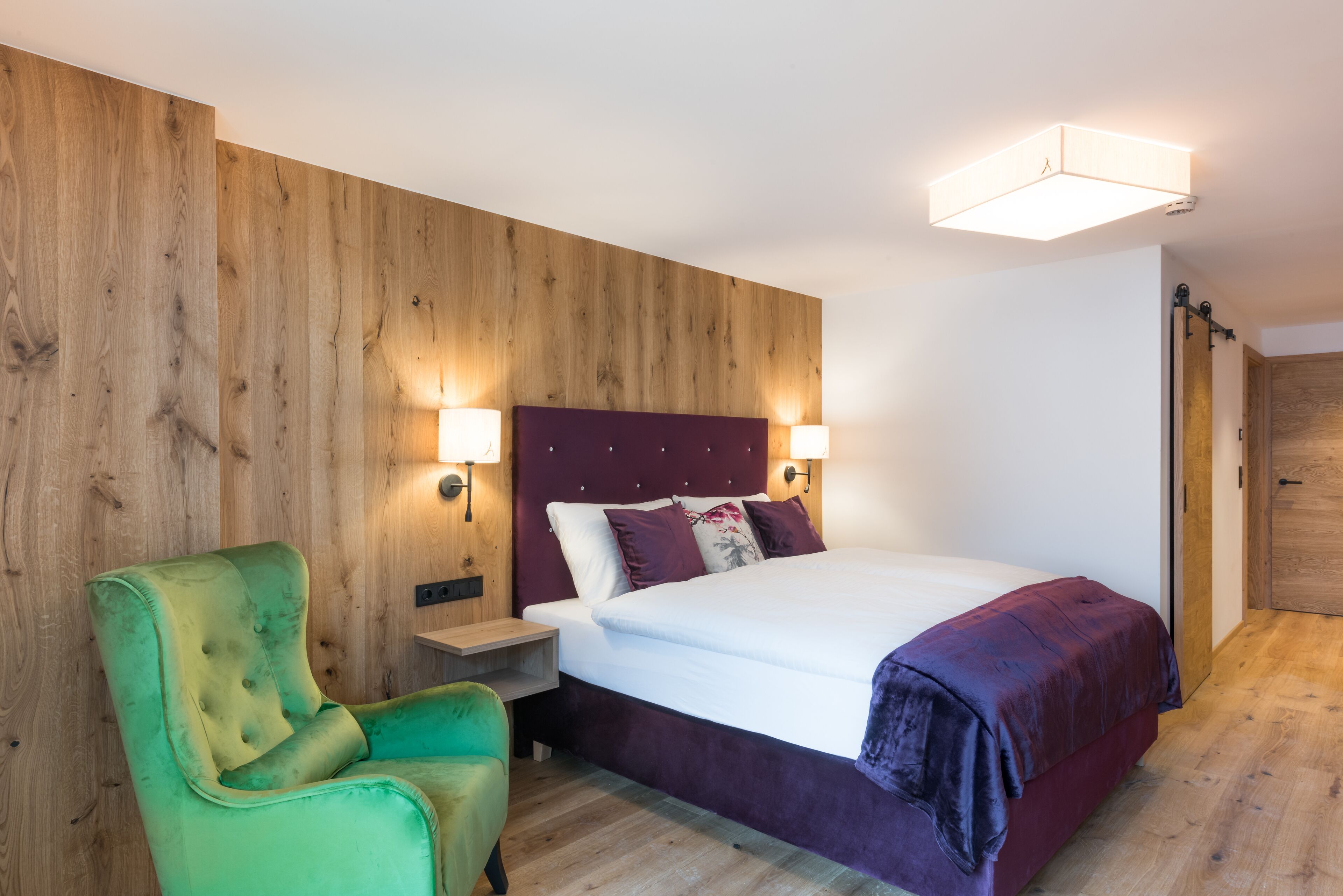 A bedroom with a wooden wall panel, a green armchair, a bed with a purple headboard, and two bedside lamps.