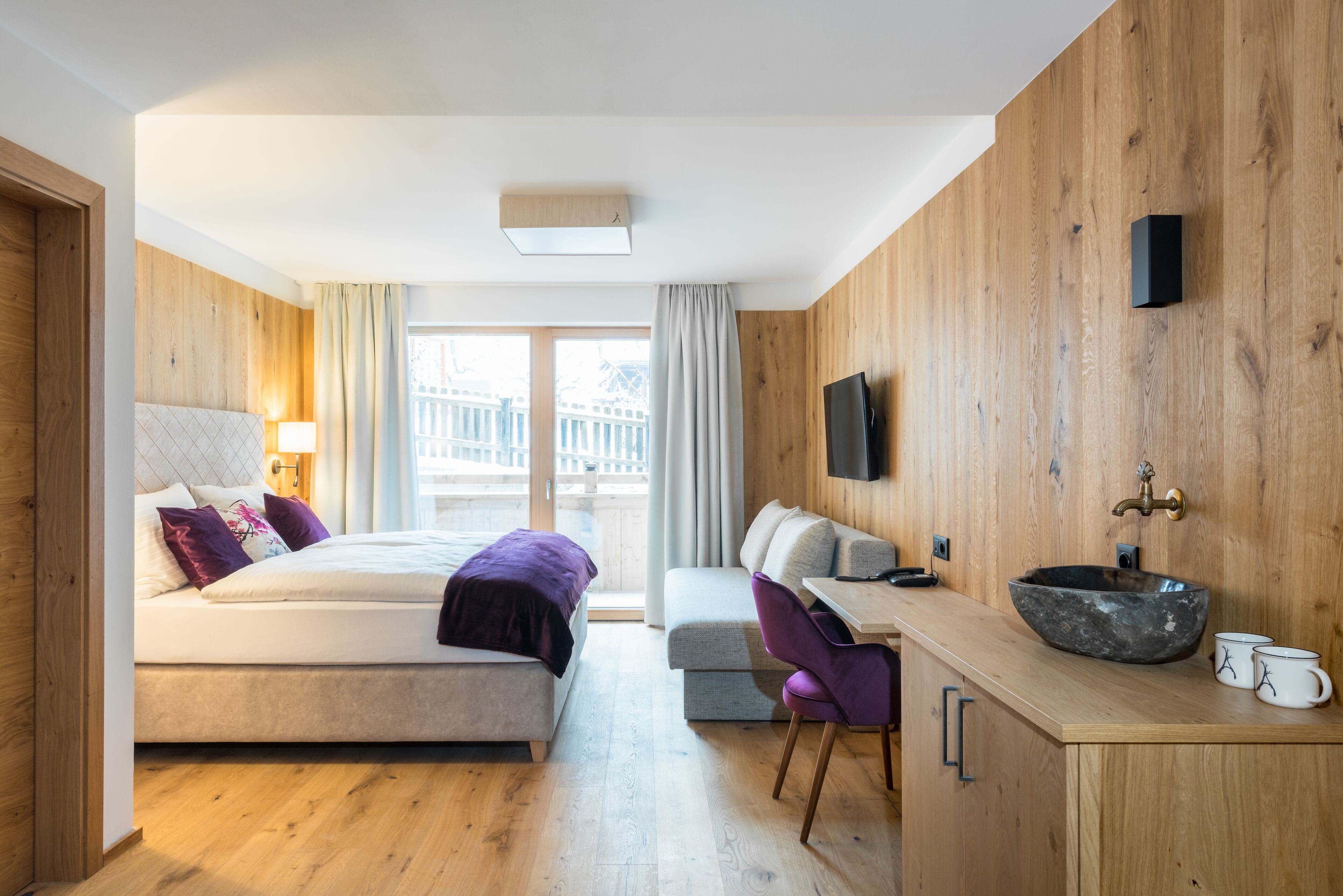 Een moderne hotelkamer met een houten vloer, een bed met hoofdeinde, een bureau met computer, een aan de muur bevestigde televisie en een raam met gordijnen.