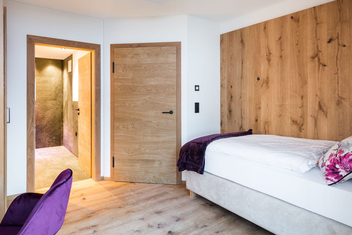 A bedroom with a wooden headboard, a bed with a white mattress and a purple blanket, a purple chair, and a wooden wardrobe.