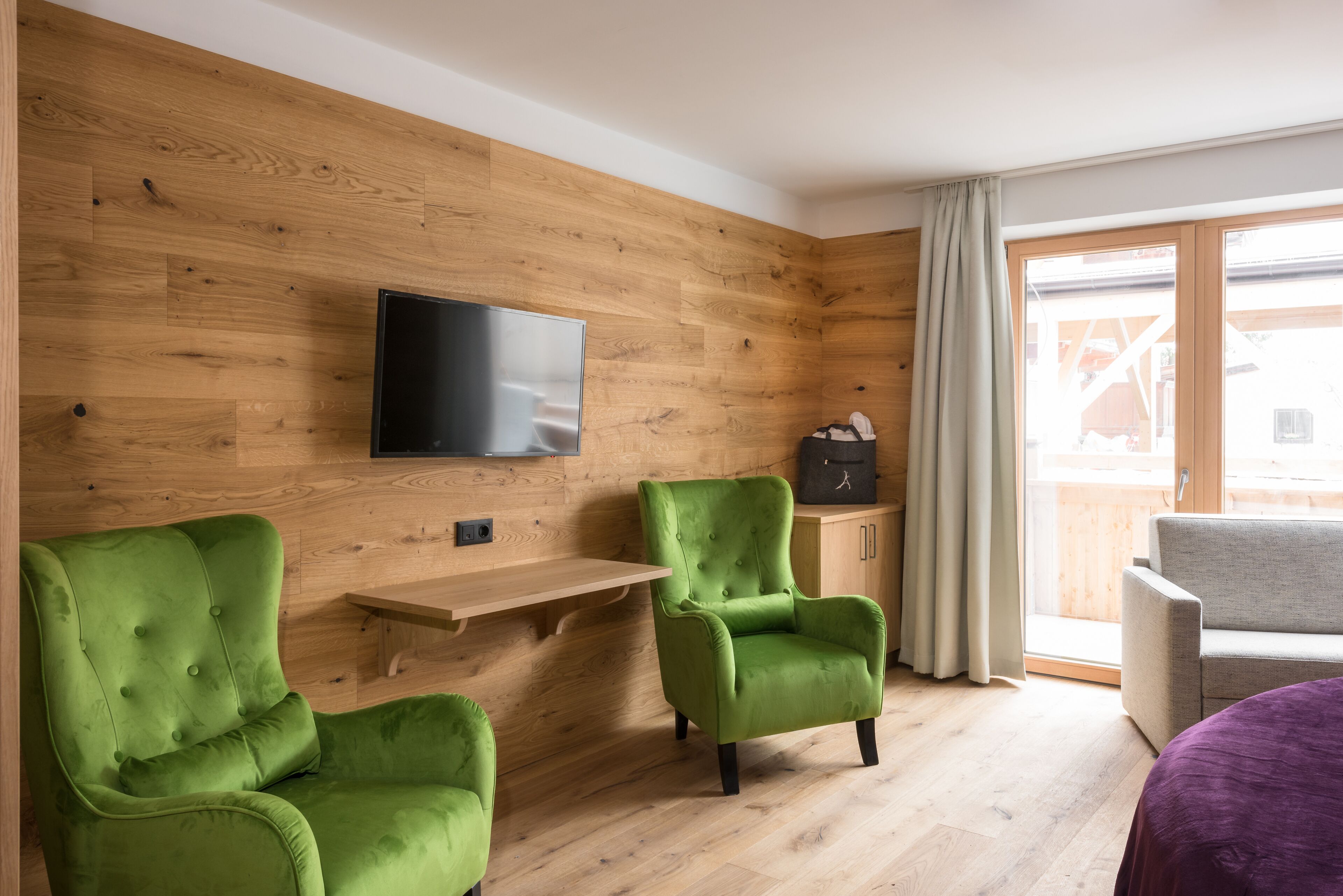 Ein modernes Wohnzimmer mit Holzwandverkleidung, einem an der Wand befestigten Flachbildfernseher, zwei grünen Sesseln, einem grauen Sofa und einer Holzbank.