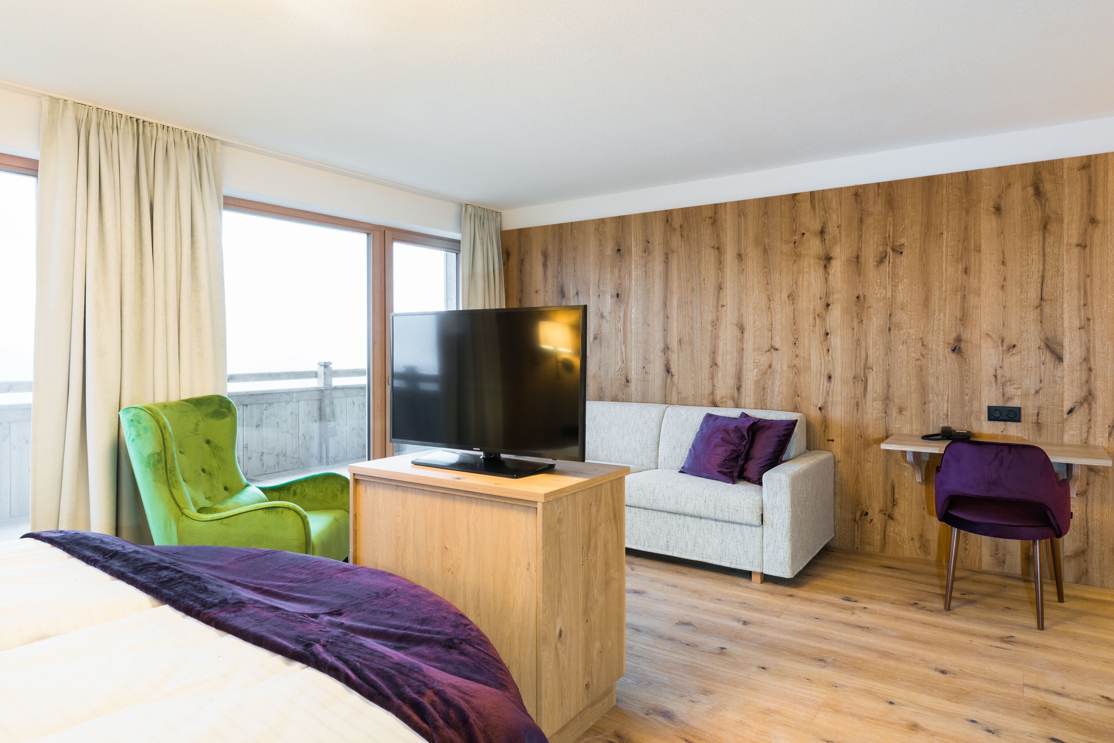 A hotel room with a bed, a green armchair, a sofa, a TV, and a wooden wall.