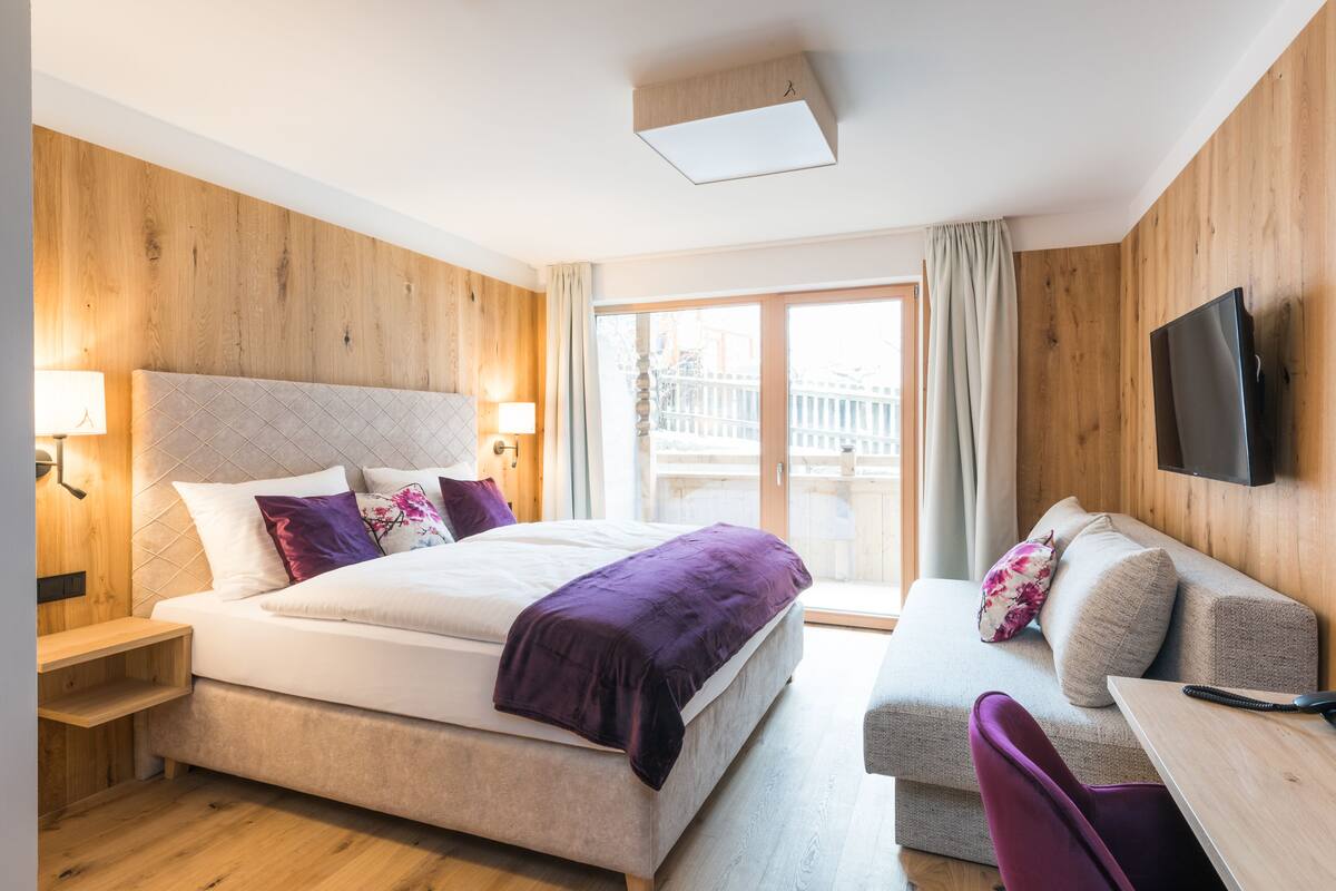 A modern hotel room with a large bed, a wooden headboard, a flat-screen TV mounted on the wall, and a balcony with a wooden railing.