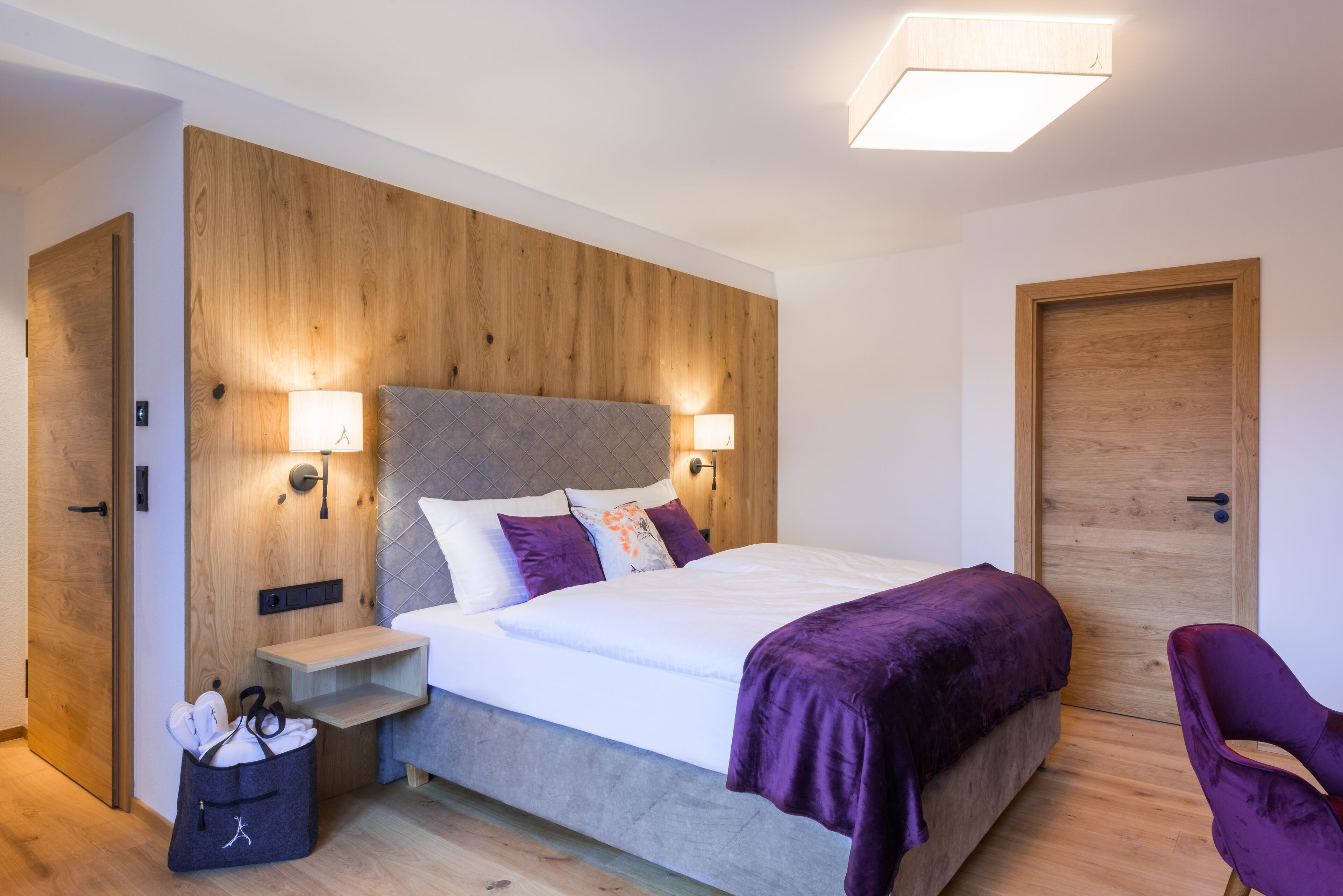 Ein modernes Schlafzimmer mit einem Holzkopfende, zwei Nachttischlampen, einem Bett mit violetter Decke, einem kleinen Nachttisch und einem Sessel.