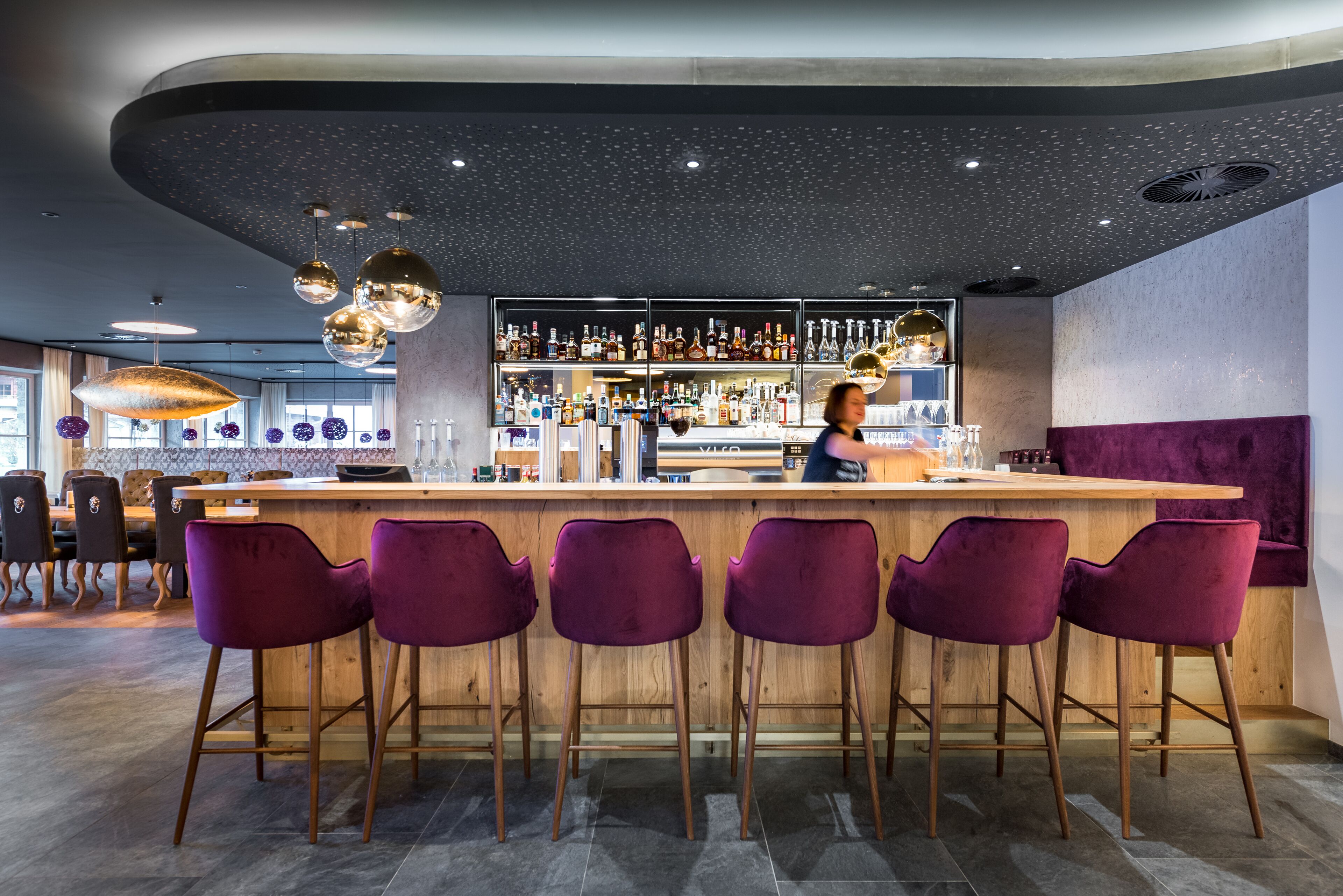 Zona de bar con mostrador de madera, taburetes morados y una variedad de botellas y vasos en estantes.