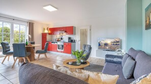 Una sala moderna con un sofá, sillas, una mesa de centro y un televisor. Se observa una cocina con armarios rojos y un comedor con una mesa y sillas.