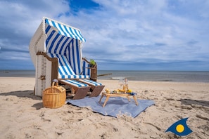 En strandloungesofa med stribede markiser, liggestole, en fletkurv og et bord med drikkevarer og frugt.