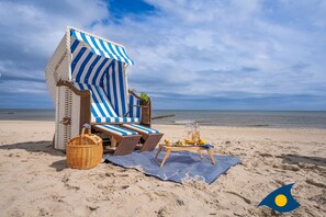 Un área de descanso frente al mar con toldos a rayas, sillas de salón, una cesta de mimbre y una mesa con bebidas y frutas.