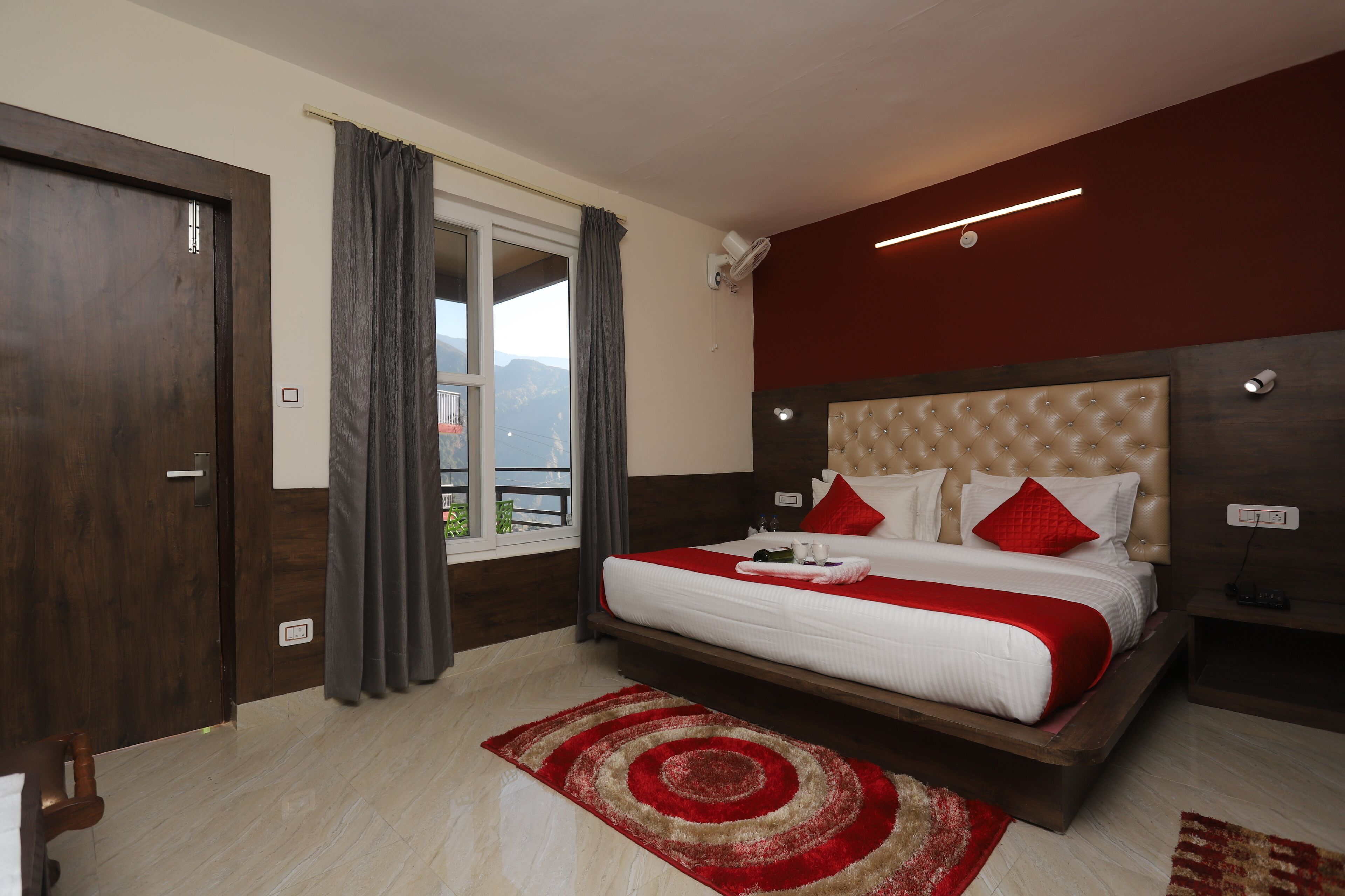 A hotel room with a large bed, a view of the mountains, a red and white bedspread, and a red and beige rug.