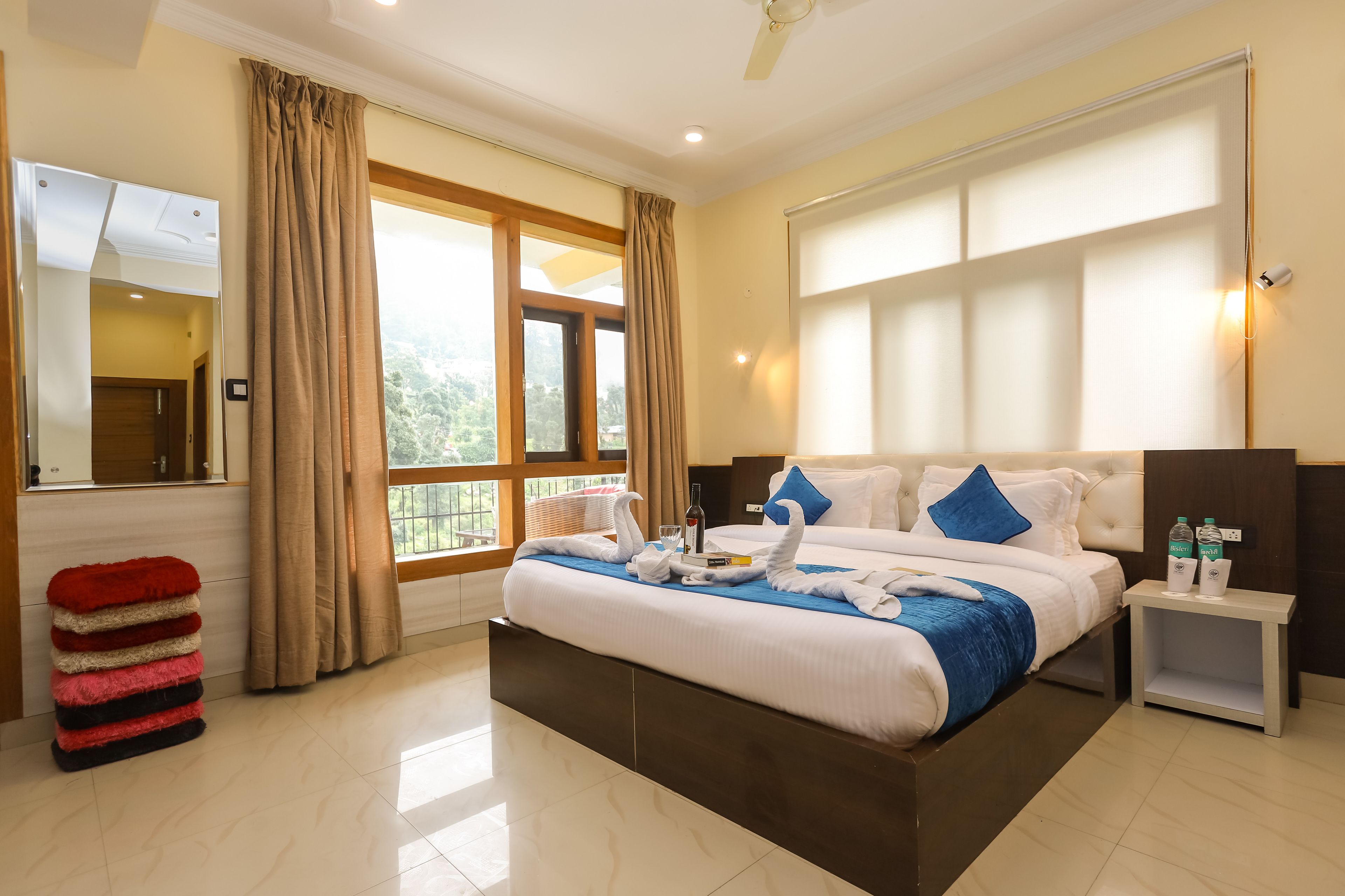 A hotel room with a large bed, a bedside table with bottles, a mirror, and a window with curtains.