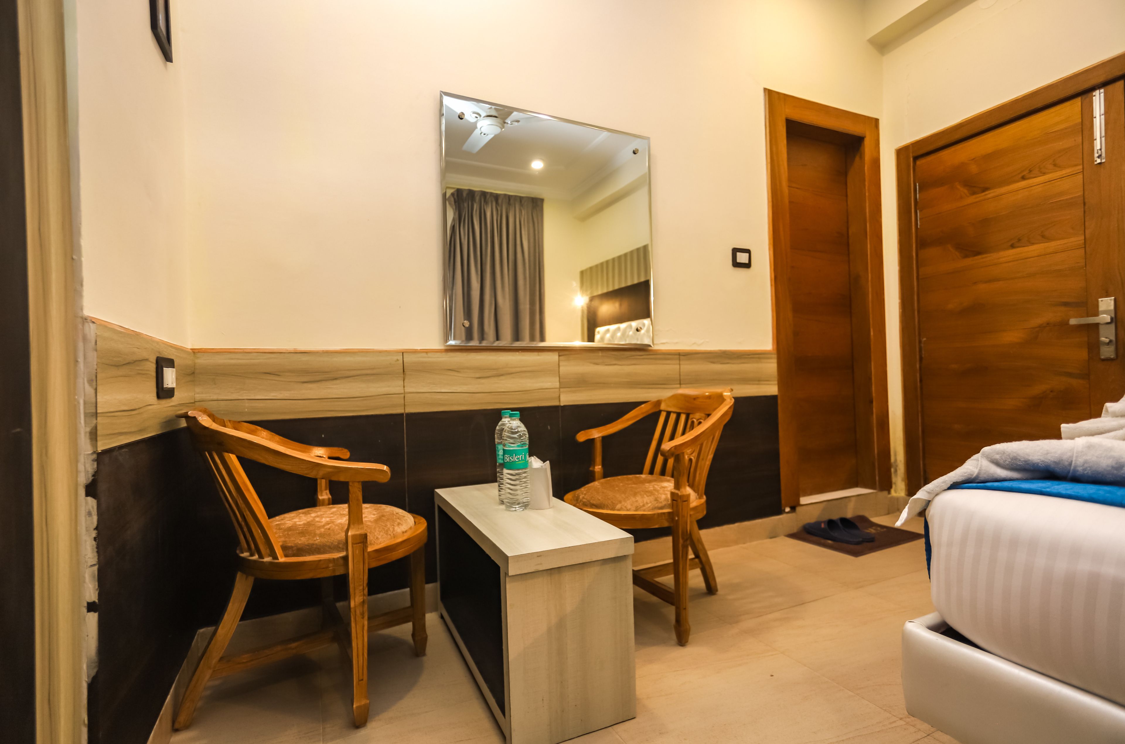 A hotel room with two wooden chairs, a small table with a bottle of water, a bed with a white comforter, a wooden door, and a mirror on the wall.