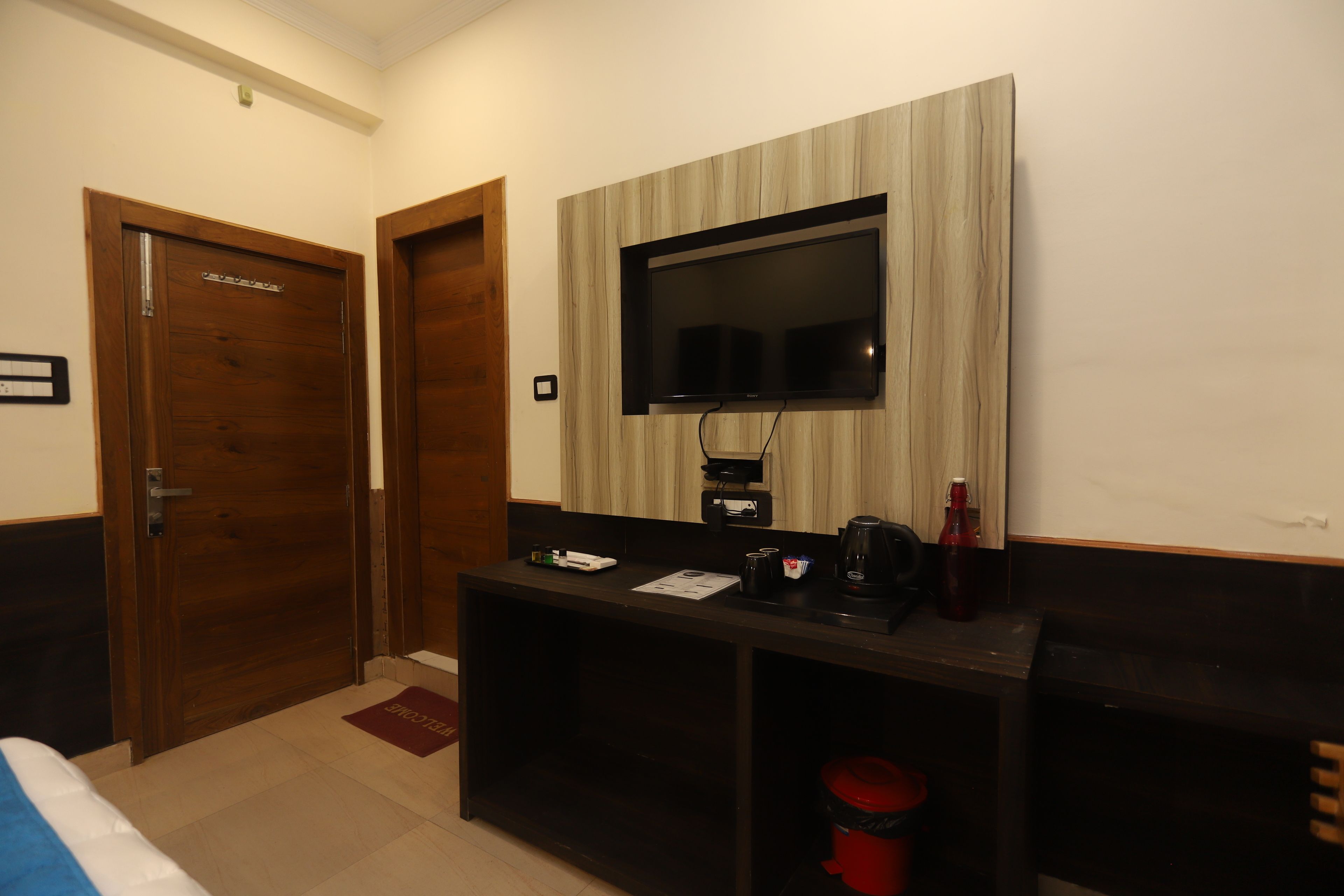 Una habitación de hotel con una puerta de madera, un televisor montado en la pared, un escritorio con cafetera y un cubo rojo.