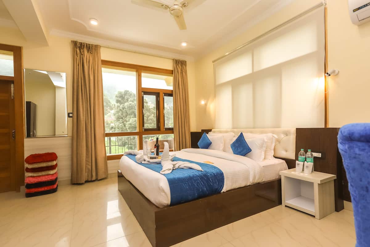 A hotel room with a large bed, a nightstand with water bottles, a red and black striped stool, and a view of trees through the window.