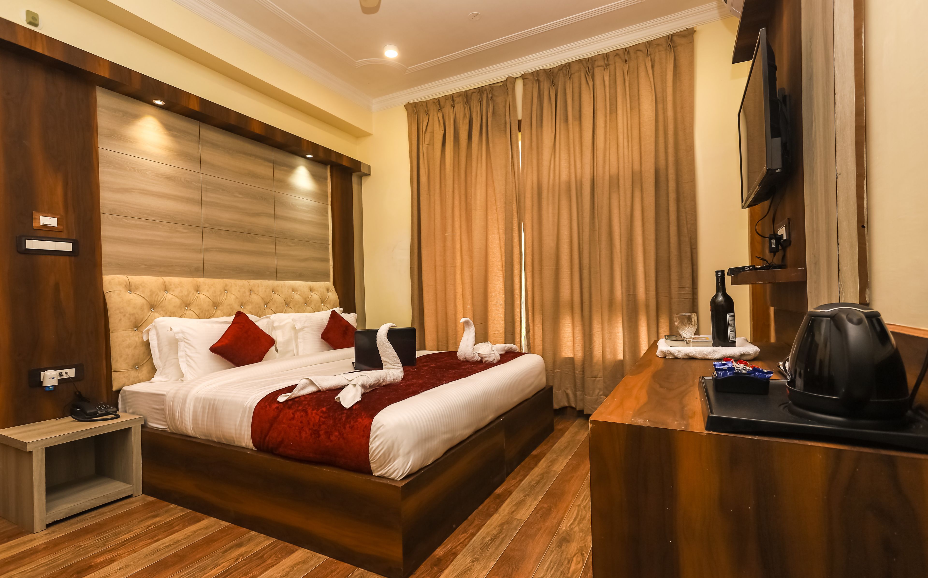 A hotel room with a large bed, a wooden nightstand, a desk with a kettle, and a television on the wall.