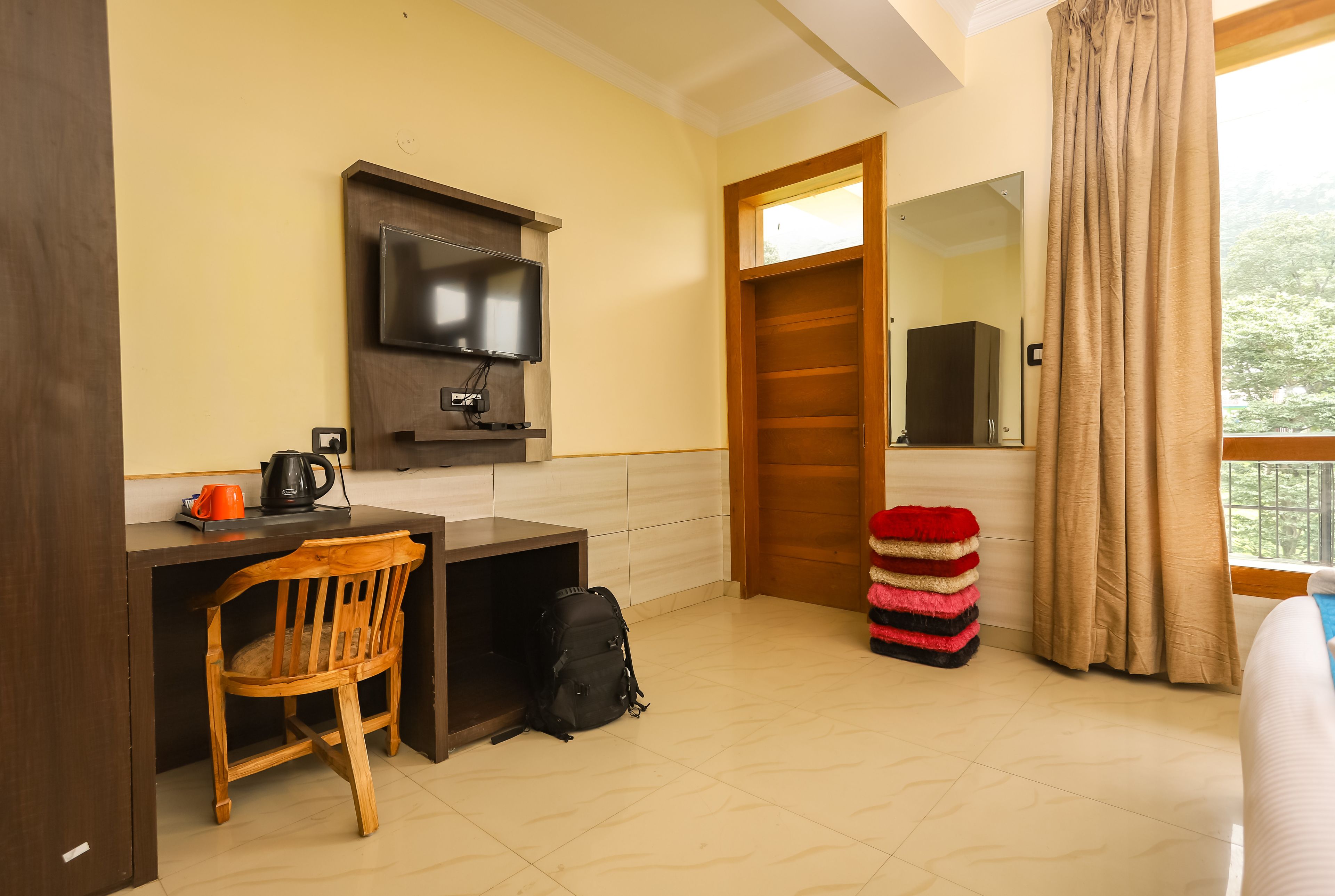 A hotel room with a wooden desk, a television, a chair, a backpack, and a window with curtains.