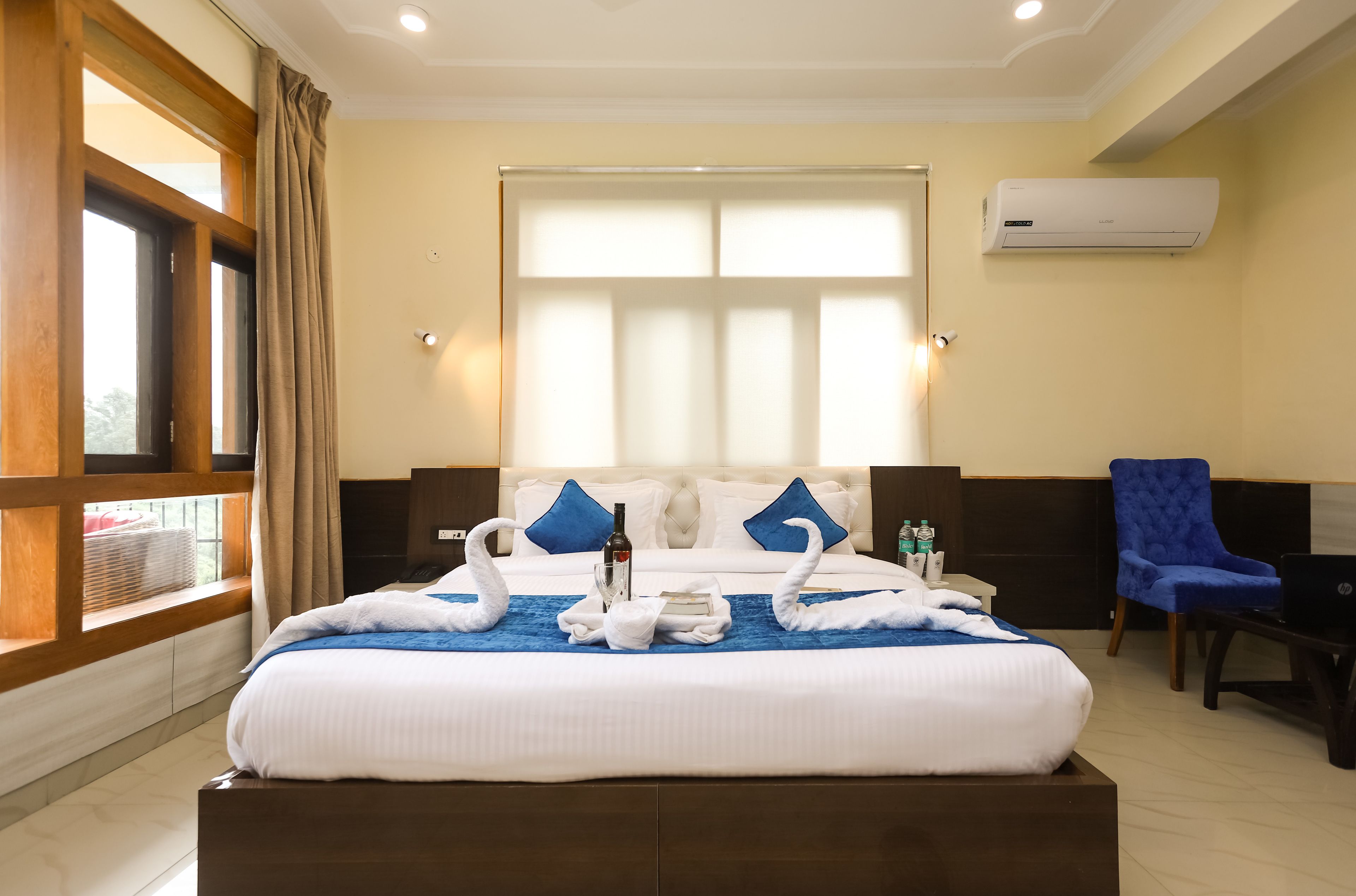 A hotel room with a large bed, two blue pillows, a window with curtains, a blue chair, a desk with a laptop, and an air conditioning unit on the wall.