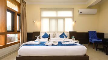 A hotel room with a large bed, two blue pillows, a window with curtains, a blue chair, a desk with a laptop, and an air conditioning unit on the wall.