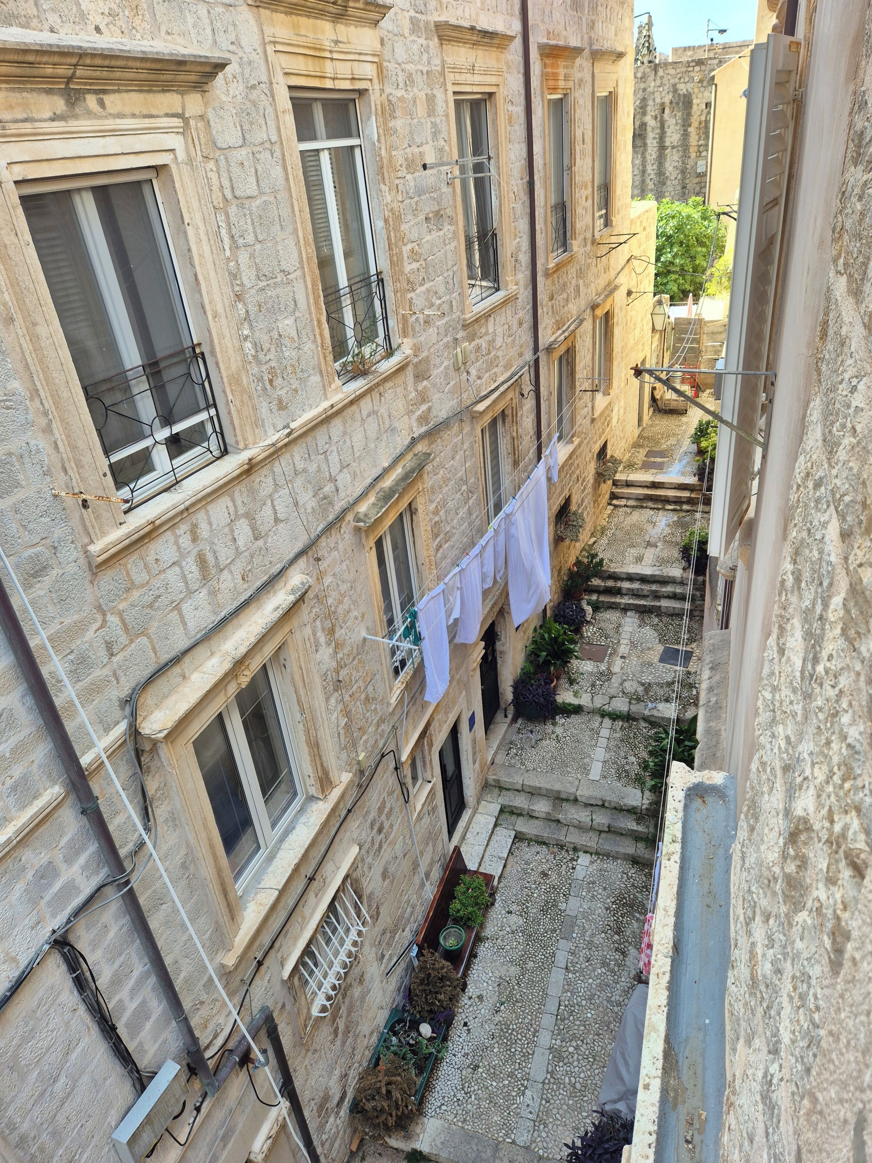 A narrow alleyway between buildings with laundry hanging on a line, a small garden area, and a visible window with a metal grate.