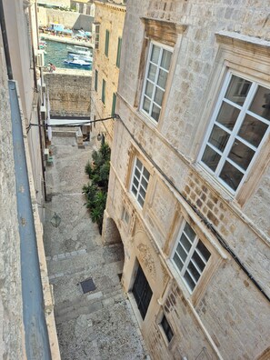Une ruelle étroite entre deux bâtiments, avec un chemin pavé de pierres, un petit espace aménagé en jardin et, au loin, une vue sur des bateaux.