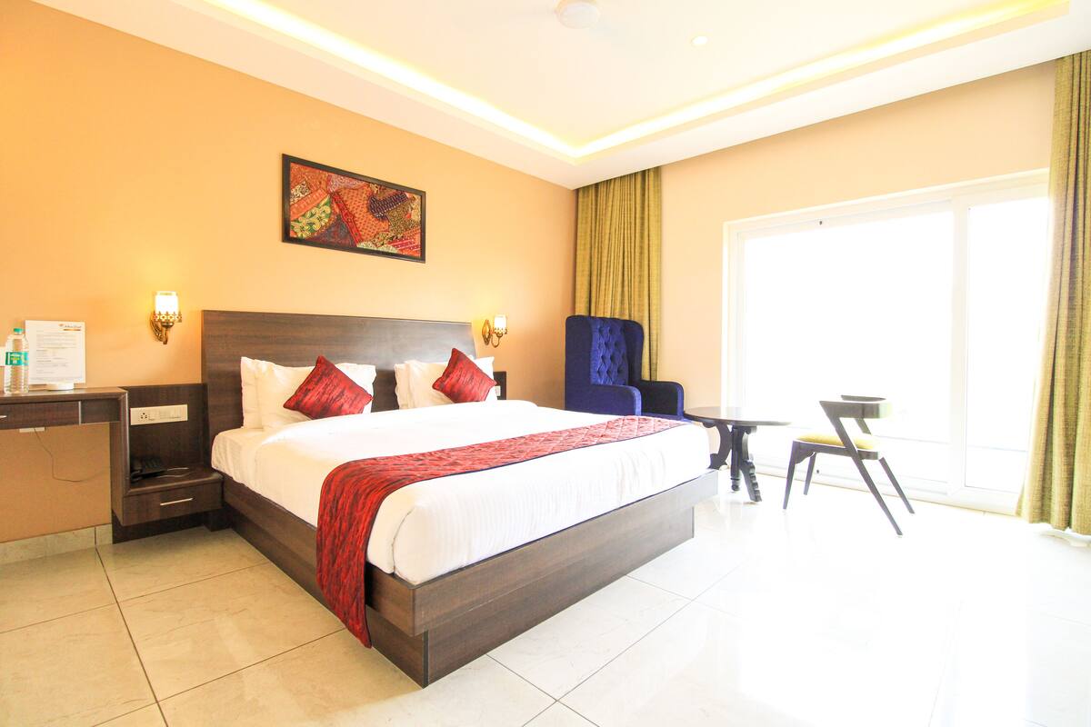 A hotel room with a large bed, bedside tables, a desk, a chair, a small table, and a large window.