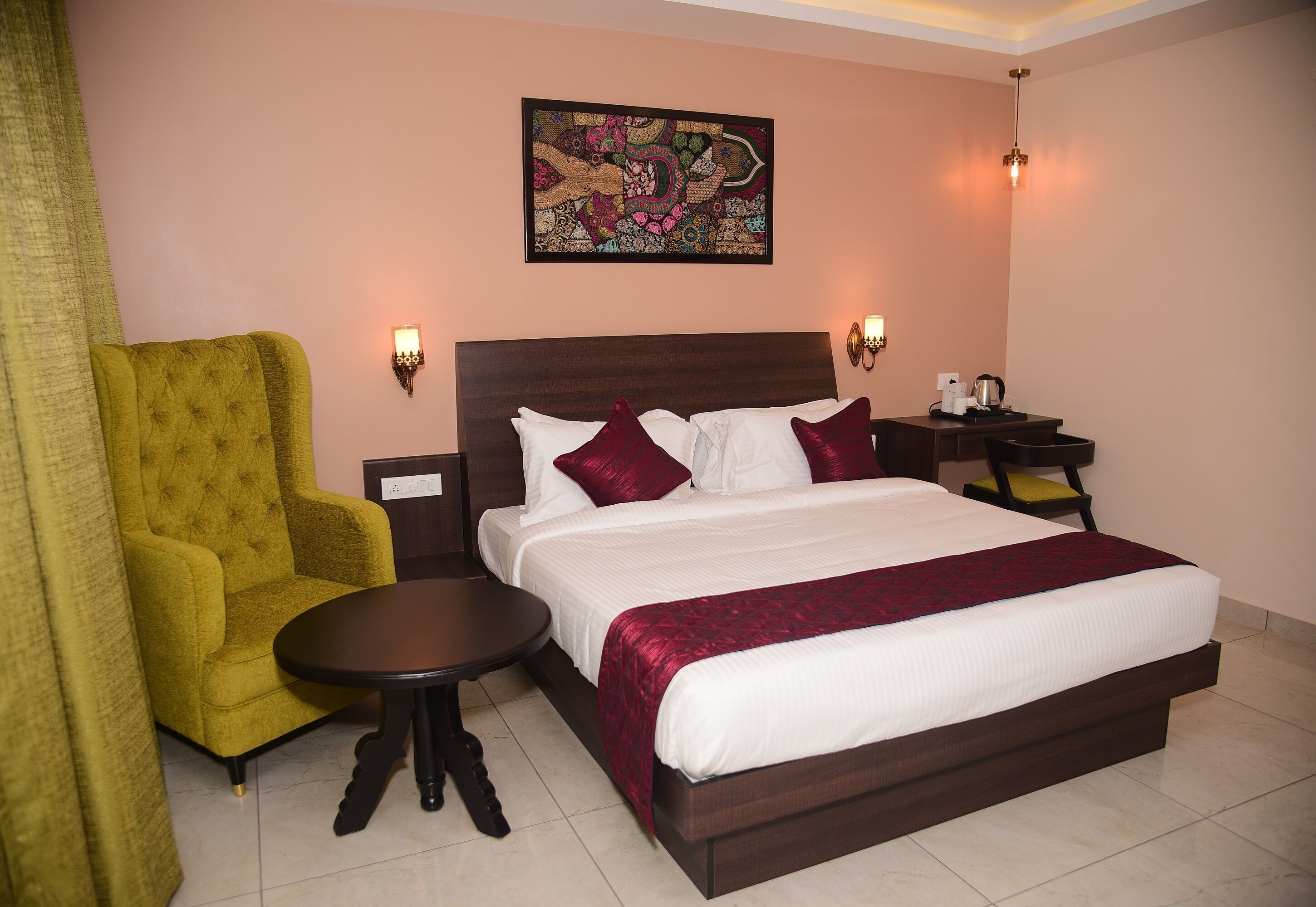 A hotel room with a large bed, a small round table, a green armchair, two bedside lamps, and a decorative wall piece.
