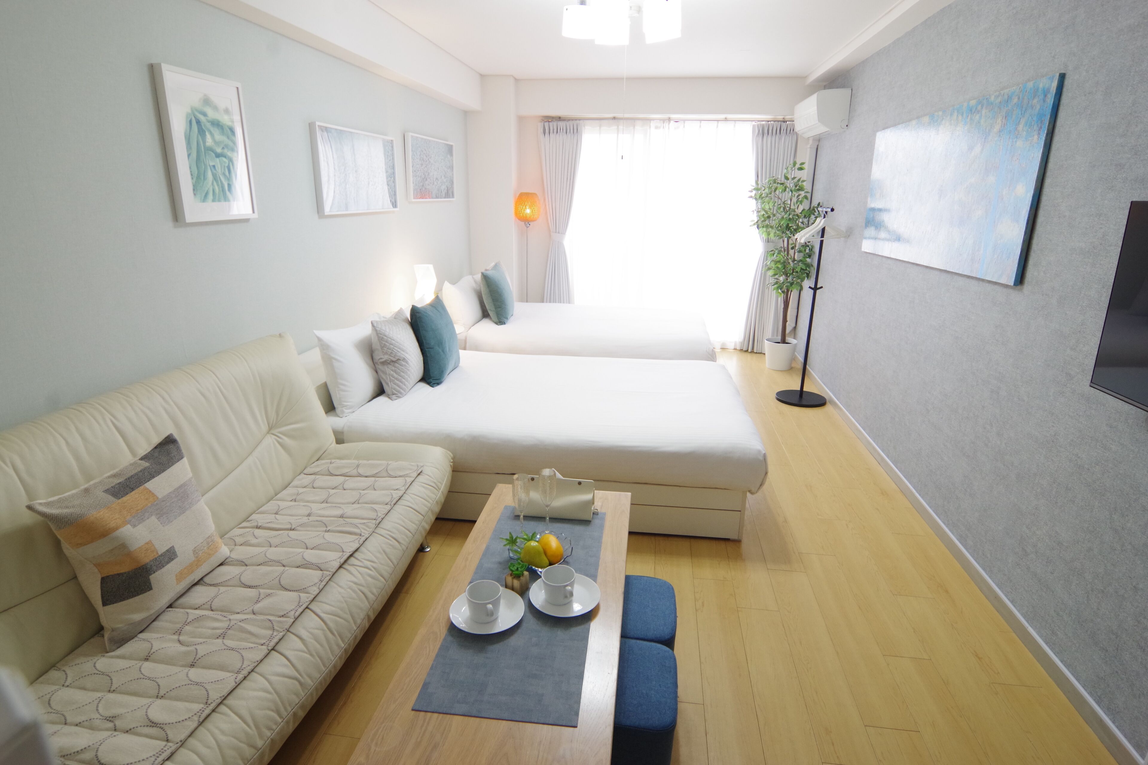 A modern hotel room with a sofa, a bed, a coffee table with cups and fruit, and a TV on the wall.