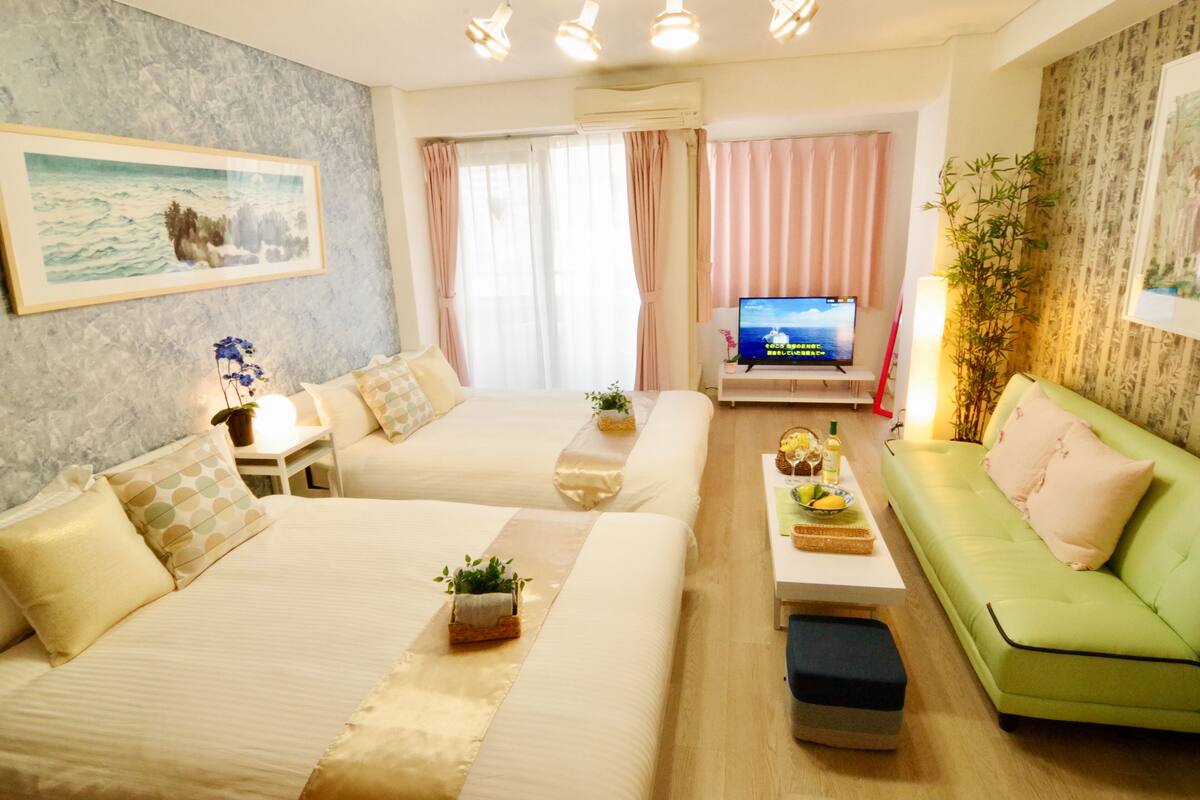 A hotel room with two beds, a green sofa, a TV, and a small table with a lamp.