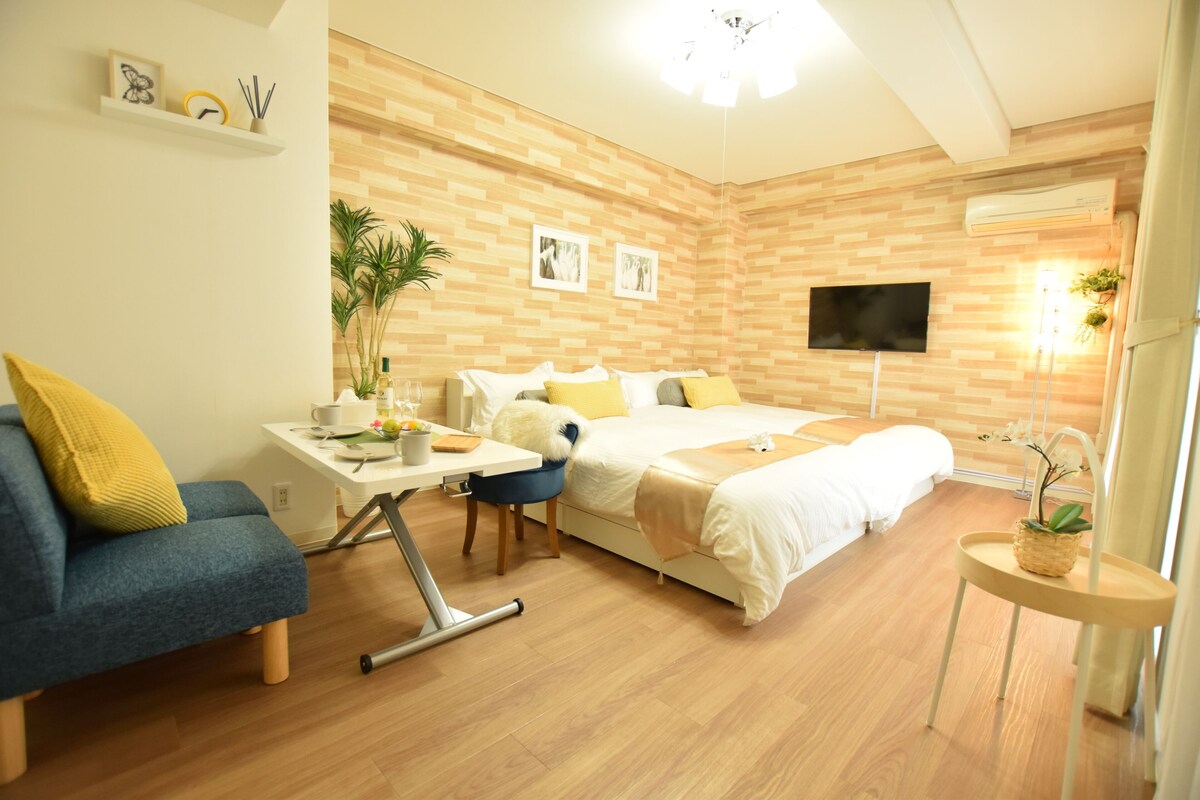 A modern hotel room with a wooden floor, a bed with white and yellow bedding, a desk with a chair, a small table with a plant, and a TV mounted on the wall.
