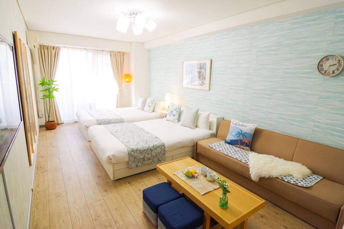 A hotel room with two beds, a sofa, a table with fruits and cups, a clock, and a wall with a textured pattern.