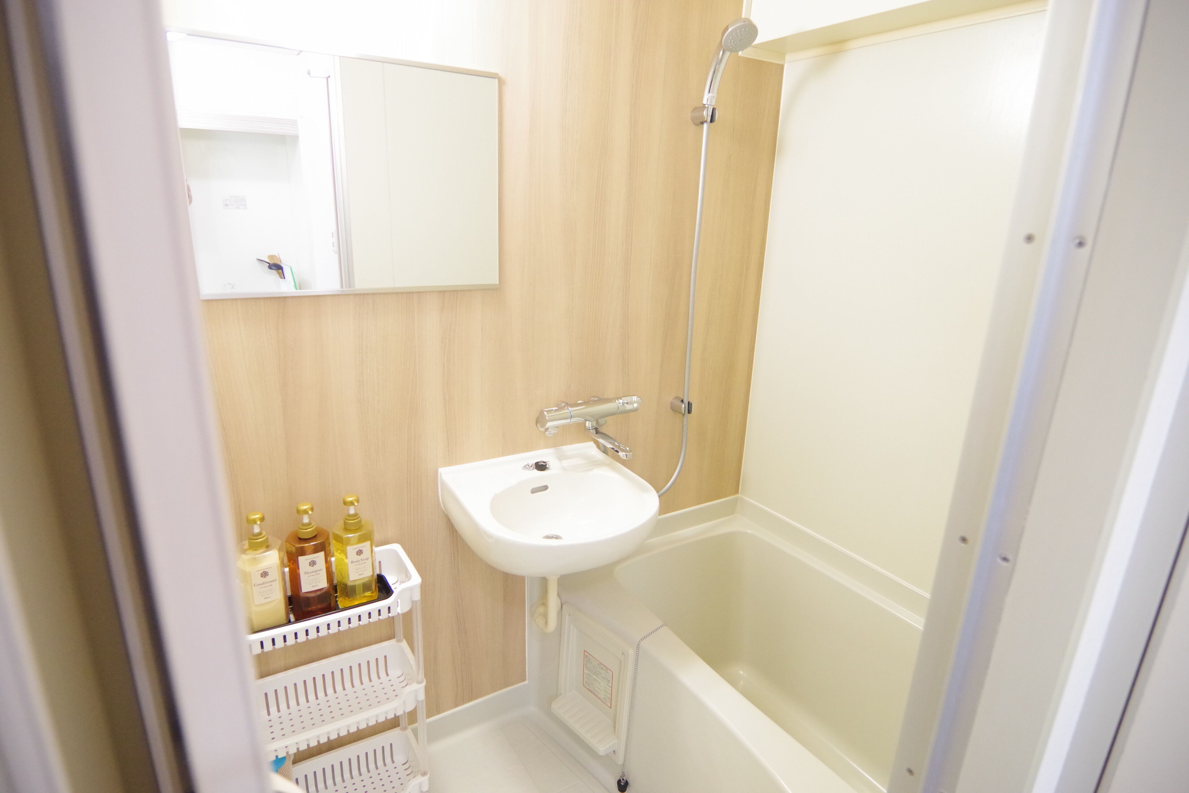 Baño con lavamanos blanco, ducha y espejo. Hay botellas en una repisa.