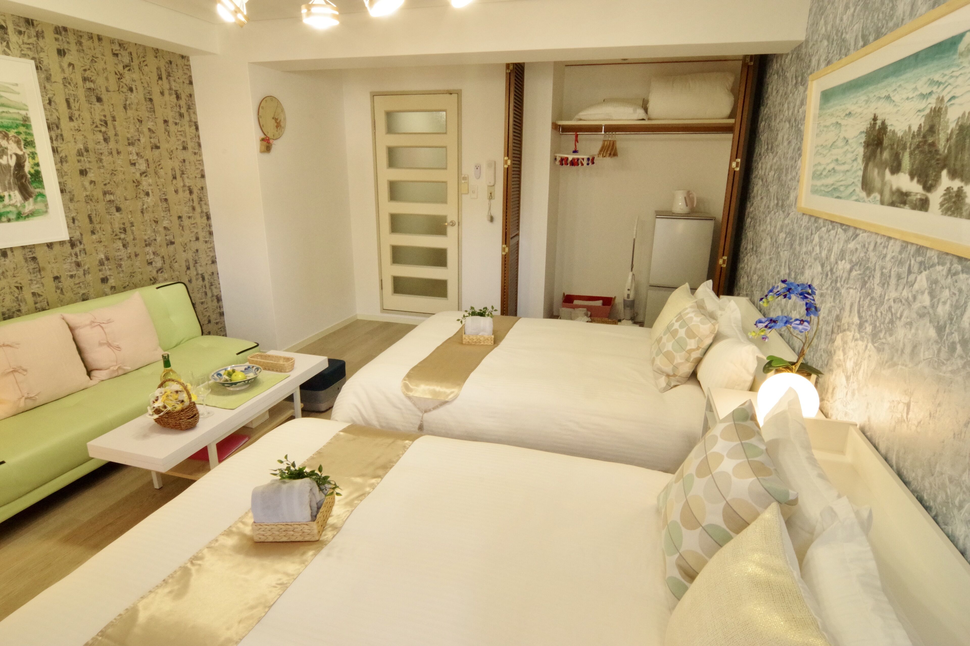 A hotel room with a green sofa, two beds with white linens, a small table with a basket and a bottle, and a painting on the wall.