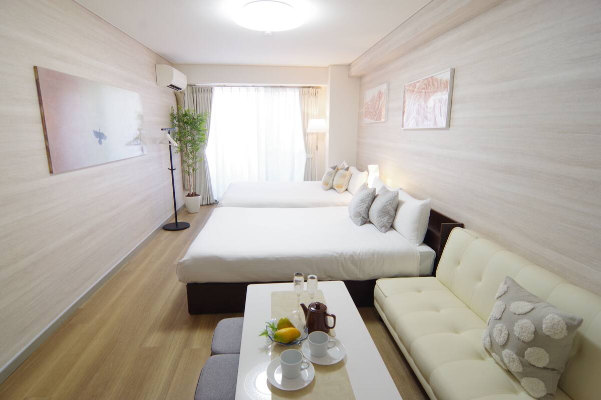 A hotel room with a bed, a sofa, a table with a pitcher, cups, and a bowl of fruit.