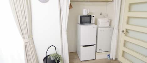 Habitación funcional con refrigerador blanco, microondas y estante pequeño con artículos de tocador. Una silla negra con una planta está apoyada en la pared, y se ve una puerta blanca con panel de vidrio satinado.
