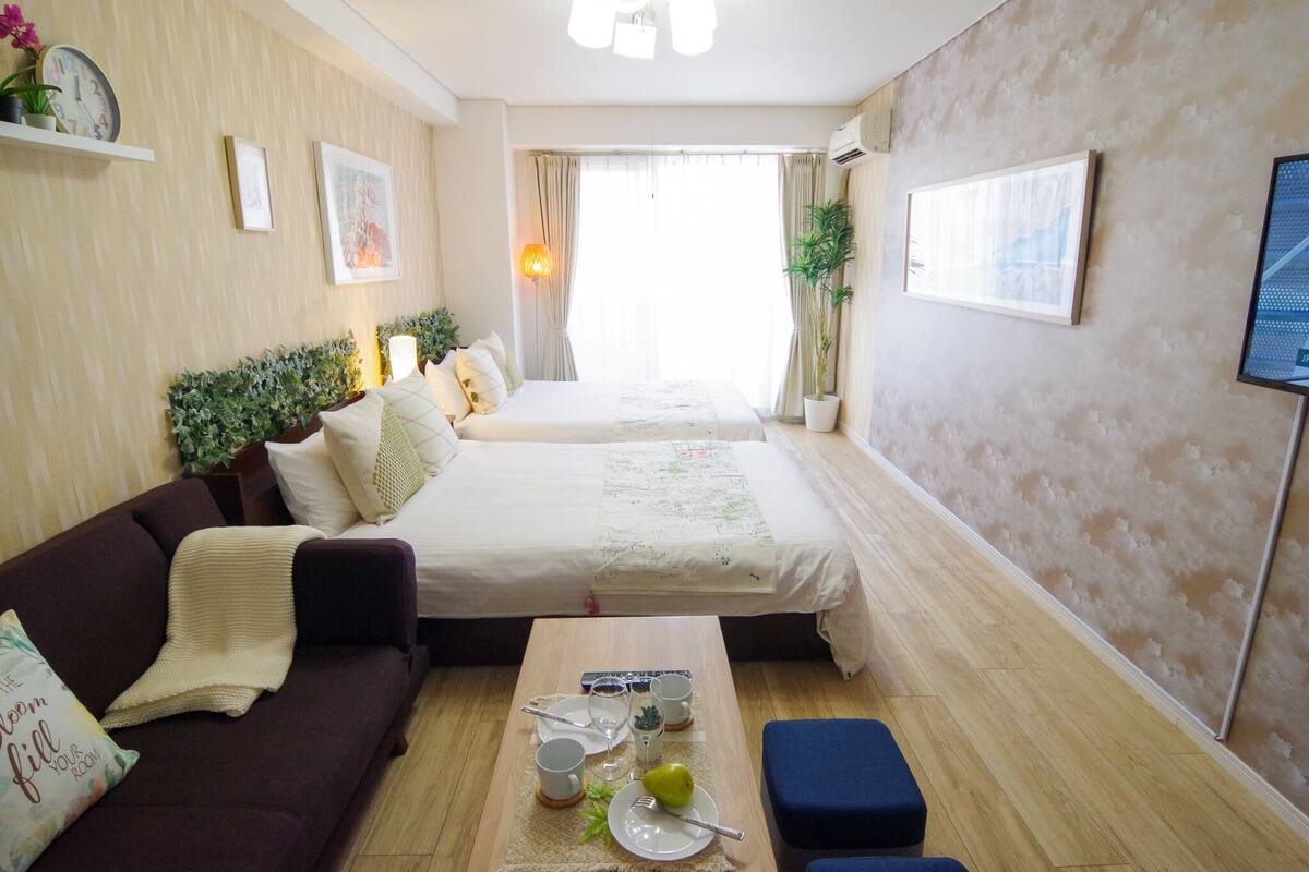 A hotel room with two beds, a sofa, a small table with a fruit and cups, and a TV on the wall.