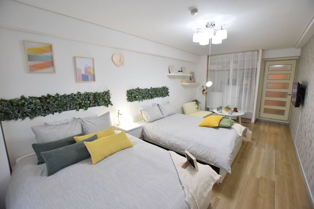 A hotel room with two beds, a bedside table with a lamp, a small round table with a plant, and a TV on the wall.