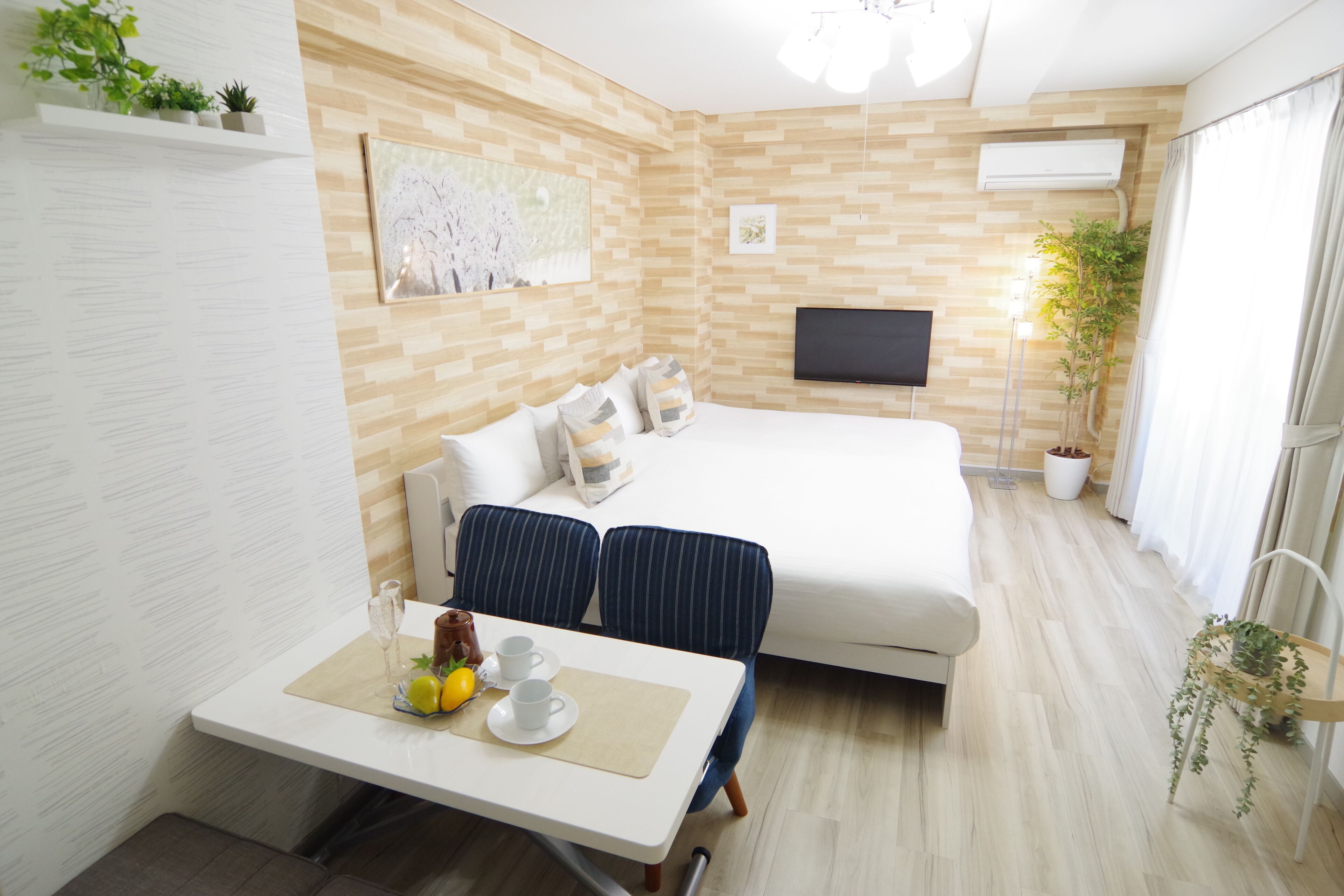 A modern hotel room with a bed, a TV, a dining table with a plant, and a window with curtains.