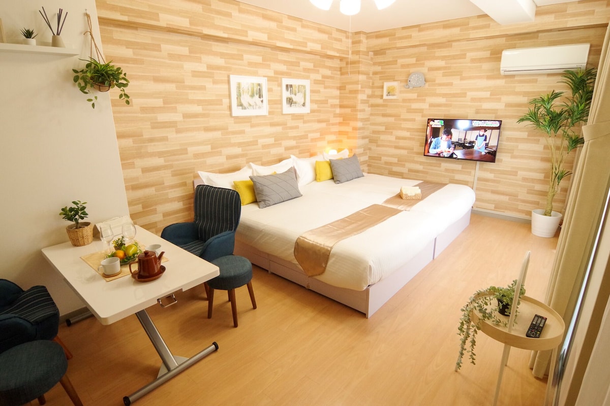 A hotel room with a bed, bedside table, chairs, a TV, and wall-mounted shelves.
